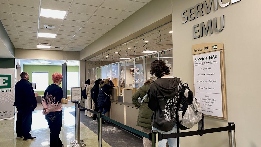 8 people wait in front of a service desk with glass panelling. 4 students gather in front of the desk, and 4 stand back and wait.
