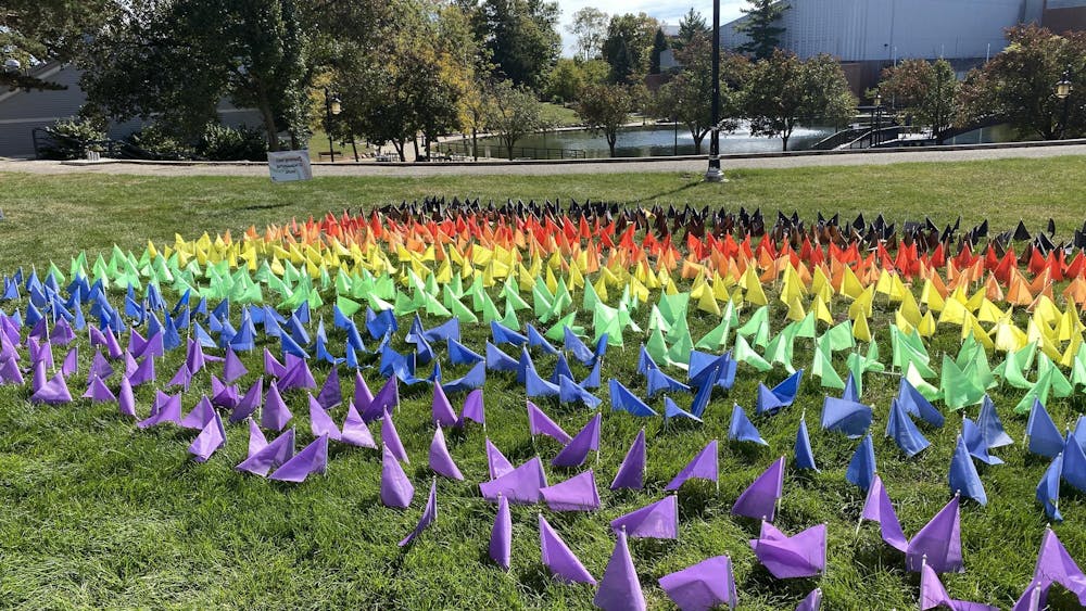 Small flags are stuck into the grass with every color of the rainbow, including black and brown, in the shape of a Pride flag.