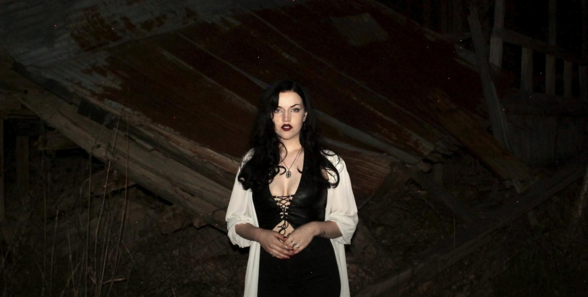 Dollie Rot, wearing a black leather dress, stands in front of an abandoned wooden structure that is falling apart.