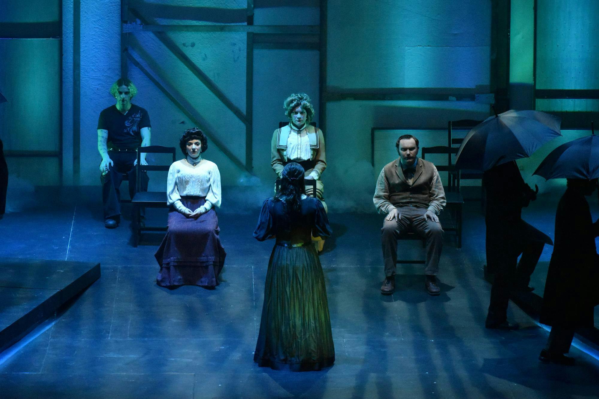 An actor stands center-stage with their back to the camera while 4 other actors sit in chairs facing them. Two actors are visible to the side of those sitting, holding umbrellas and walking across stage.