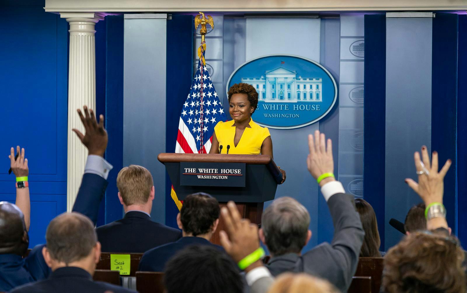 Karine Jean-Pierre stands at a White House podium in front of an American flag and a blue background and addresses a group of people with raised hands.