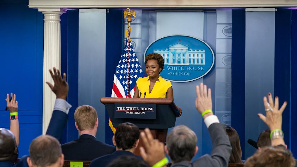 Karine Jean-Pierre stands at a White House podium in front of an American flag and a blue background and addresses a group of people with raised hands.