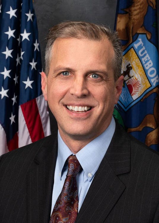 Headshot of Jeff Irwin wearing a suit with flags in the background.