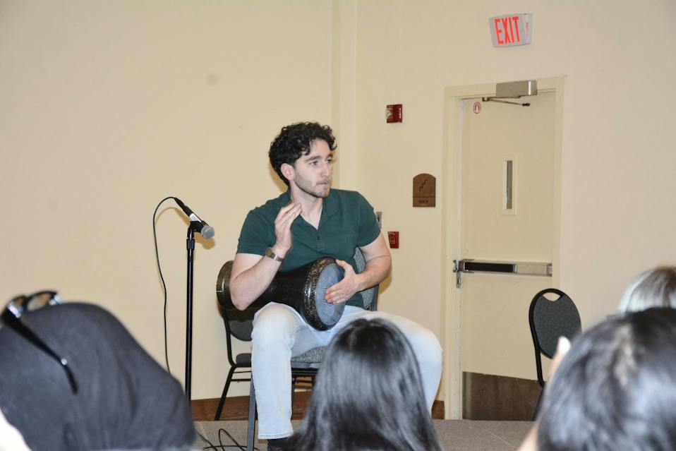 A student taps on a drum while sitting in front of a microphone.