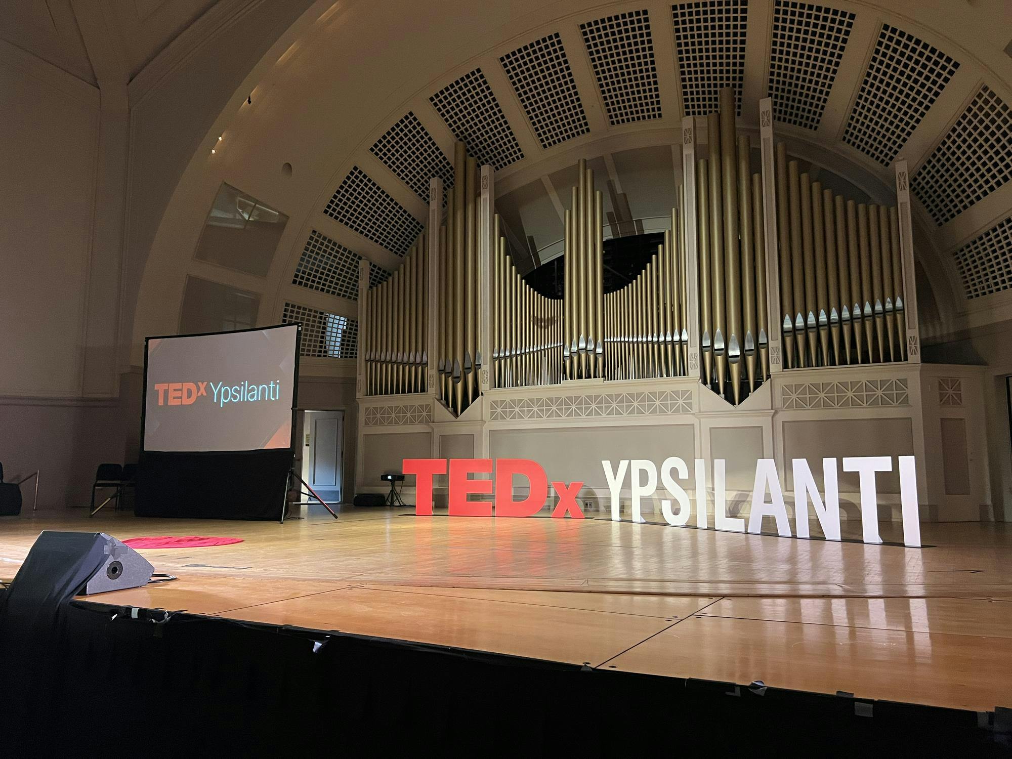 Stage lights illuminate the wooden Pease Auditorium stage. The red and white TEDxYpsilanti logo is physically stood up on stage and also displayed on a projector. A small, red circle carpet is in the middle of the stage for speakers to stand on.