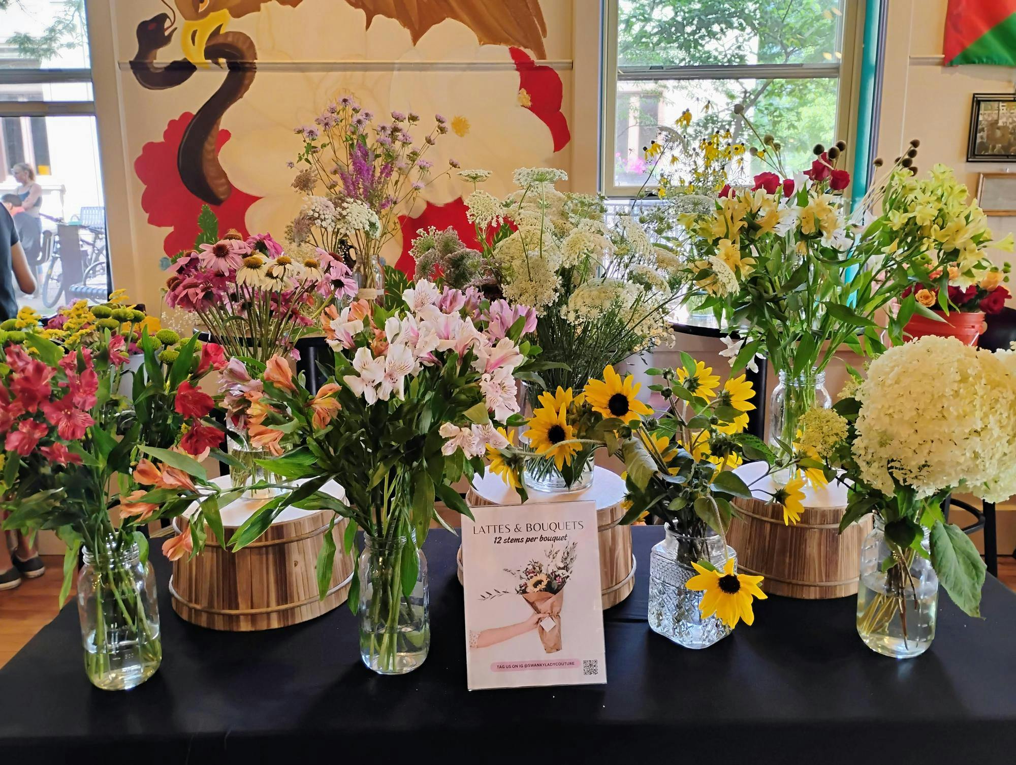 A long table with a black tablecloth is set up in the center of Bridge Community Café, and adorned with eight glass vases containing colorful cut flowers of different varieties.