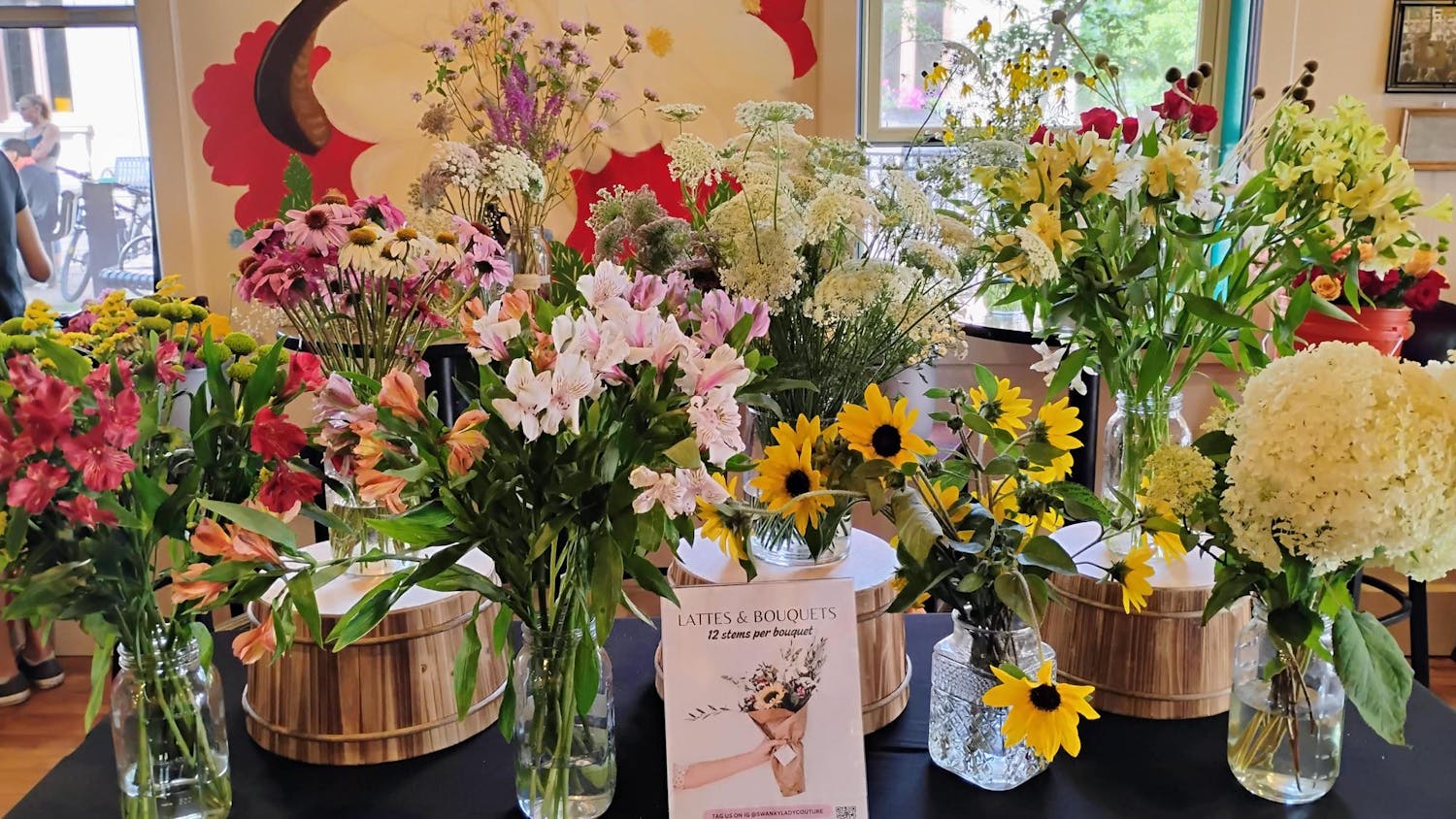 A long table with a black tablecloth is set up in the center of Bridge Community Café, and adorned with eight glass vases containing colorful cut flowers of different varieties.
