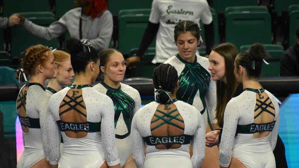 Seven EMU gymnasts huddle in a circle, listening to their coach.