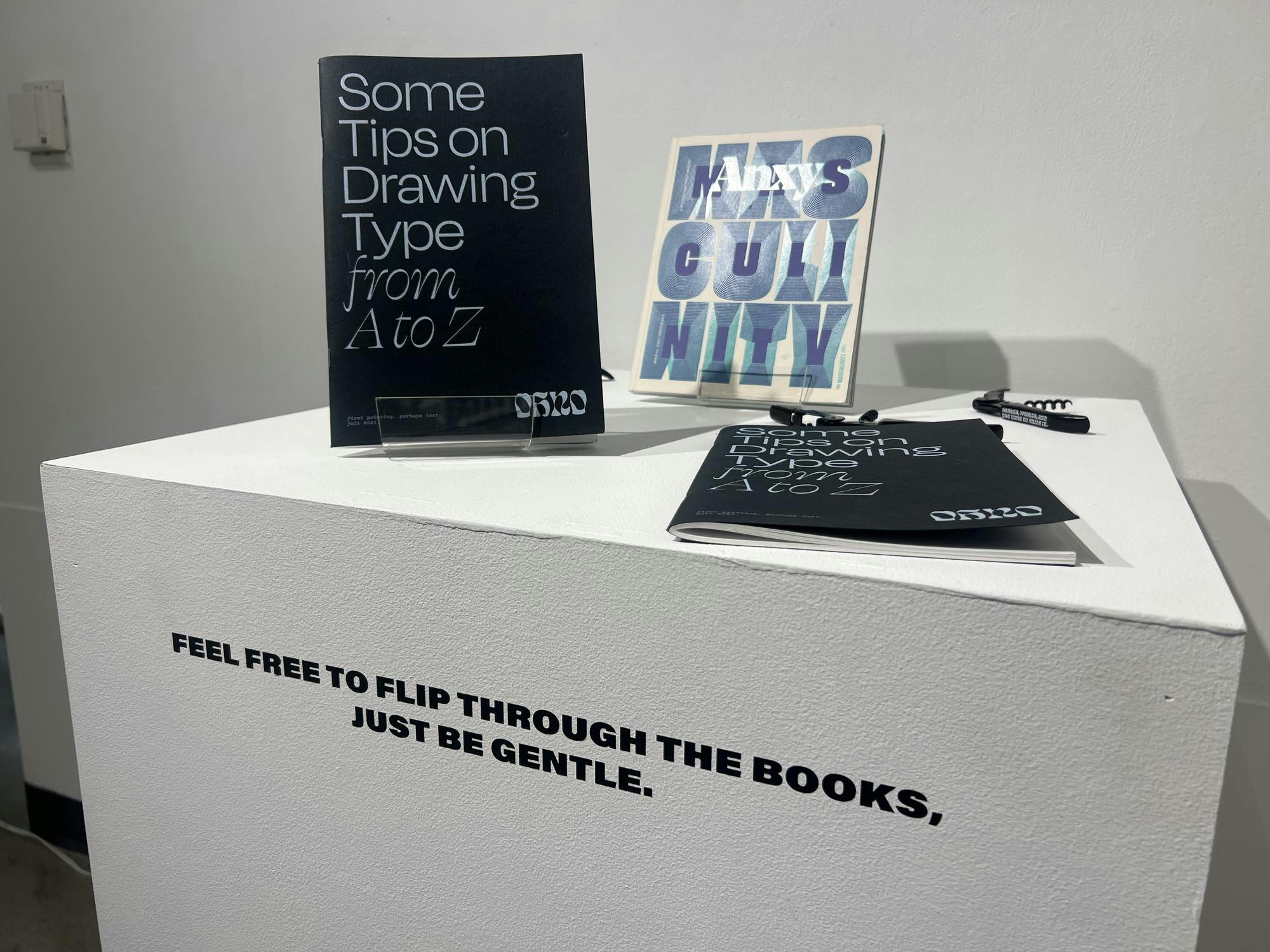 Two copies of the book "Some Tips on Drawing from A to Z" are on a white podium, along with the masculinity edition of Anxy Magazine.