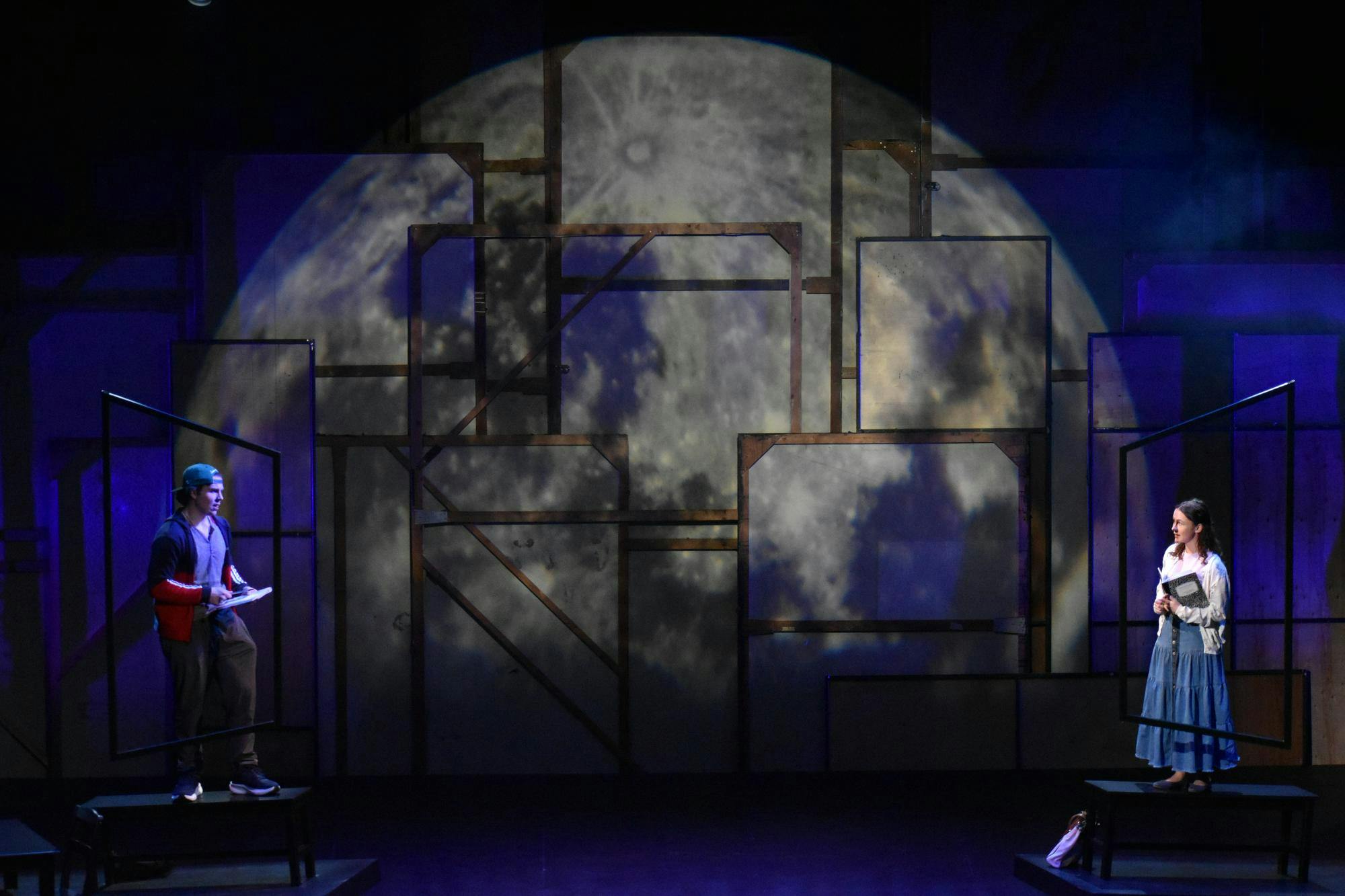 Two actors stand at far ends of the stage, looking at each other with a moon projection behind them.