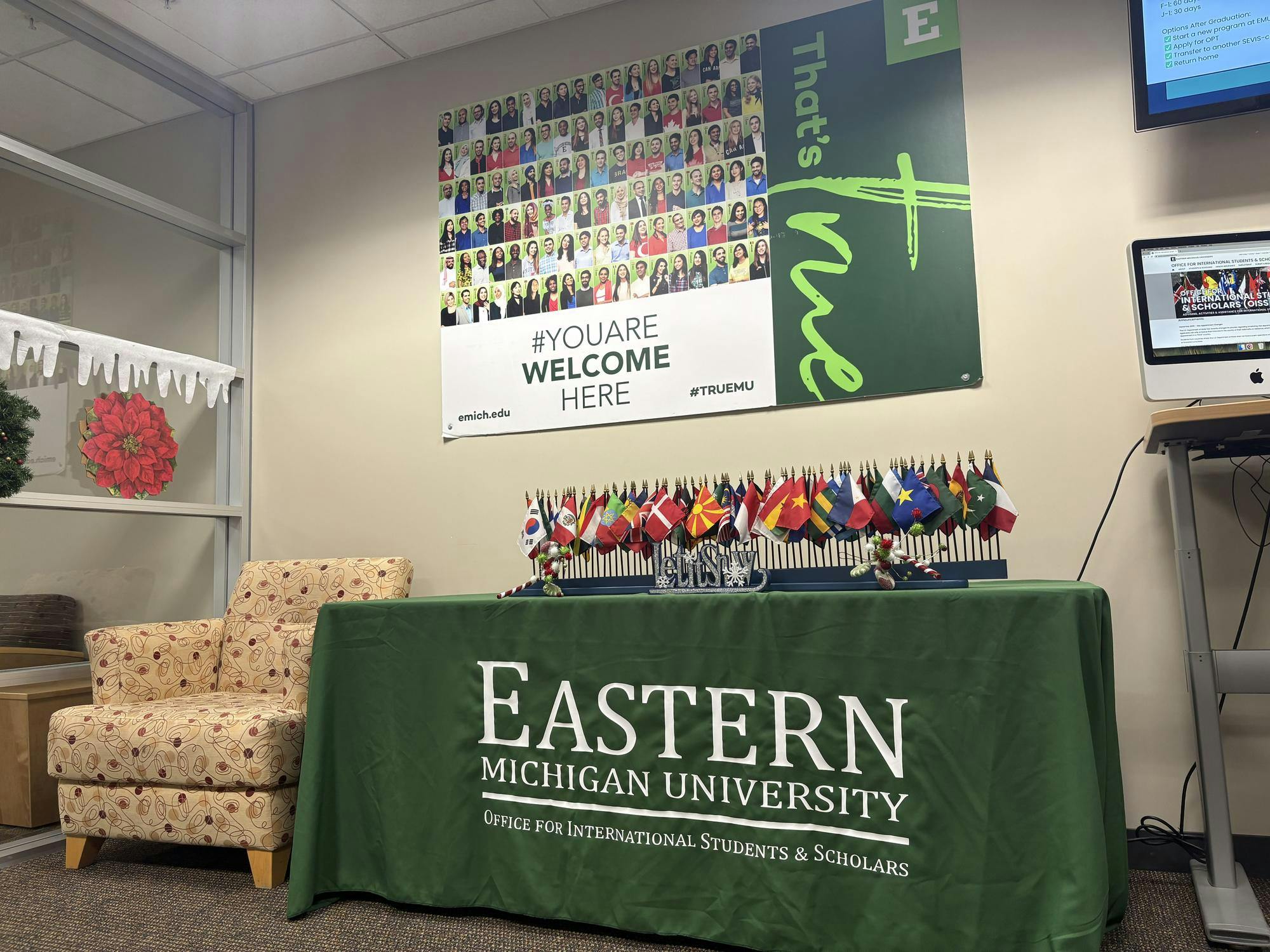 Image of a table with a green table cloth that says &quot;Eastern Michigan University Office for International Students and Scholars&quot; with a display of small flags from countries around the world. Above the table is an EMU-branded sign that says &quot;You are welcome here,&quot; with a collage of student faces.