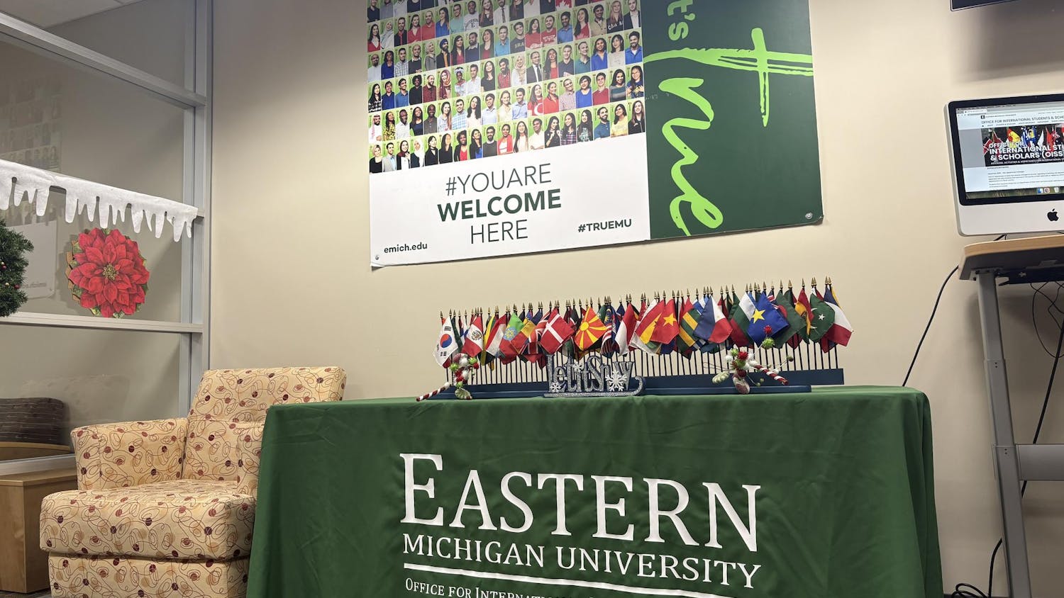 Image of a table with a green table cloth that says "Eastern Michigan University Office for International Students and Scholars" with a display of small flags from countries around the world. Above the table is an EMU-branded sign that says "You are welcome here," with a collage of student faces.