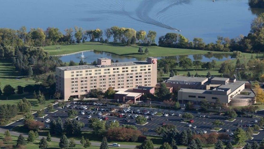 A faraway shot of a large, rectangular tan and brown building. In front of the building is a large parking lot full of cars. There are also many trees around the building and parking lot. Overhead, the sky is blue and sunny.