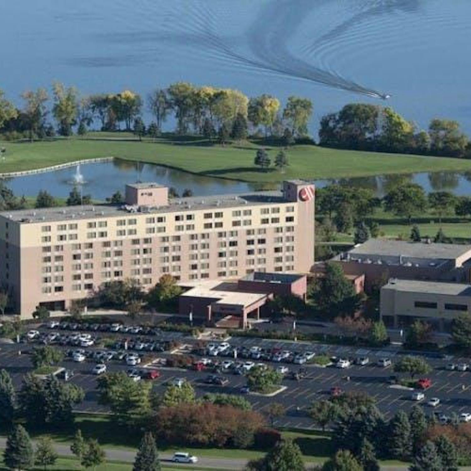 A faraway shot of a large, rectangular tan and brown building. In front of the building is a large parking lot full of cars. There are also many trees around the building and parking lot. Overhead, the sky is blue and sunny.