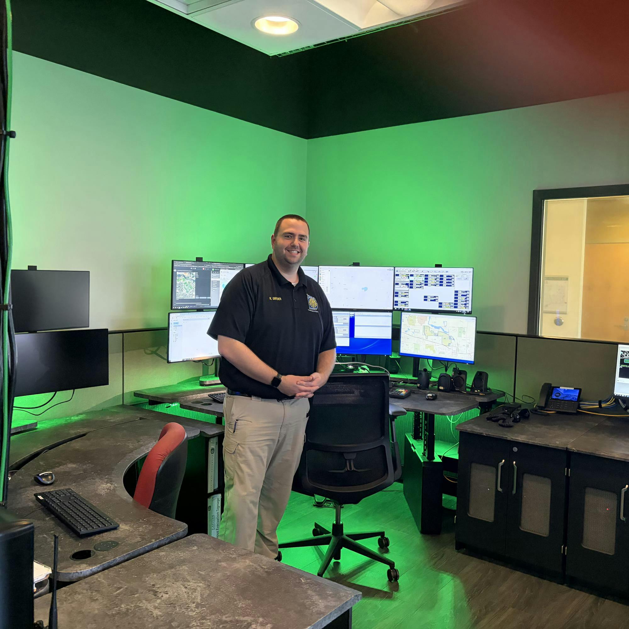 Kyle Snyder stands in front of several computer monitor screens in the dispatch center, where a glowing green light illuminates the background.