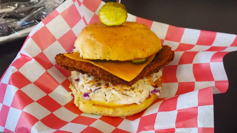 A fried chicken sandwich lies on a table. The sandwich has several layers, top to bottom: A pickle poked on top of the bun, top bun, pickles, cheddar, fried chicken, coleslaw, and a bottom bun.