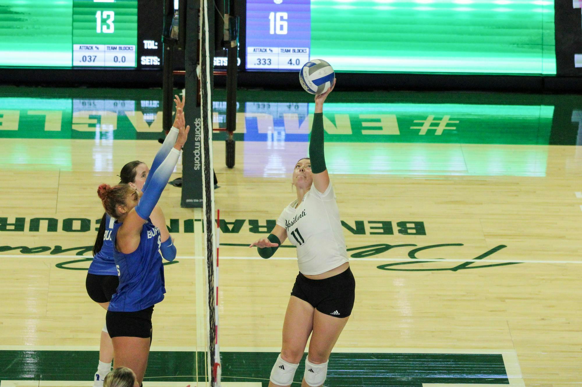 EMU Women's Volleyball player, in a white jersey, tapping the ball over the volleyball net where the Buffalo players, in blue jerseys, are waiting to block it. 