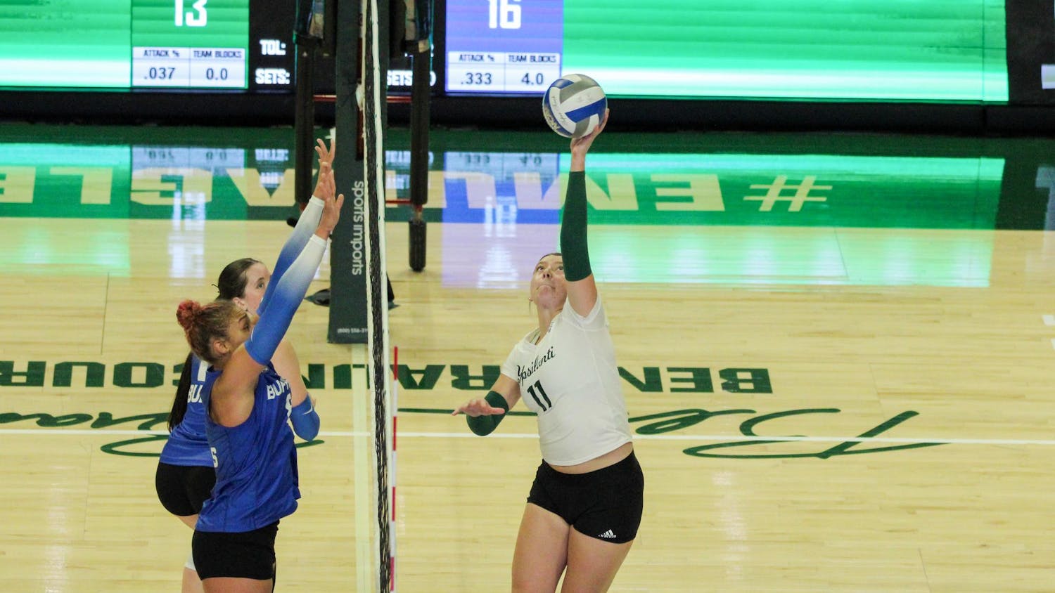 EMU Women's Volleyball player, in a white jersey, tapping the ball over the volleyball net where the Buffalo players, in blue jerseys, are waiting to block it.