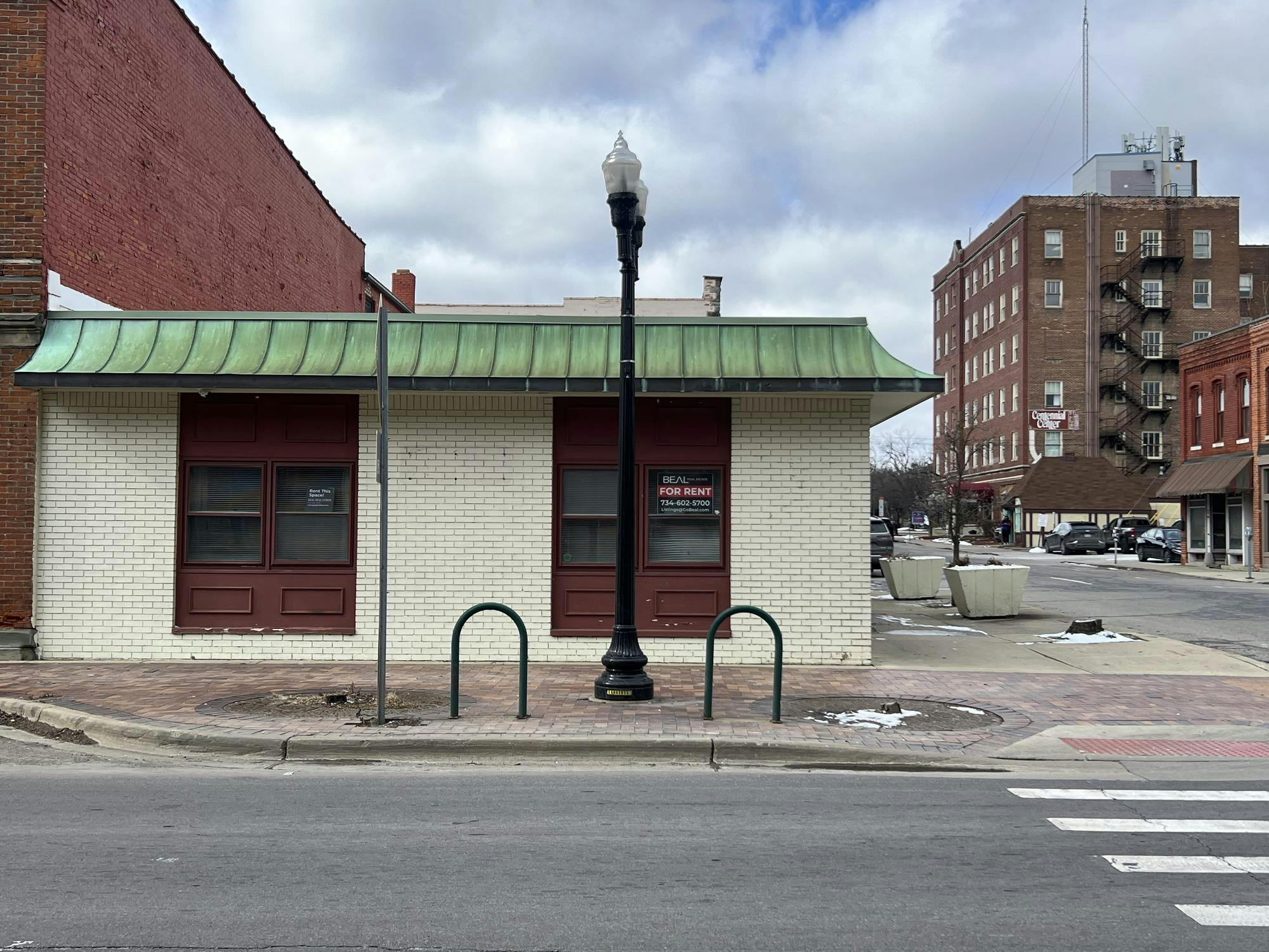 A white, brick, one-story building with red, wooden window frames. A sign in the window says that the building is available for rent.