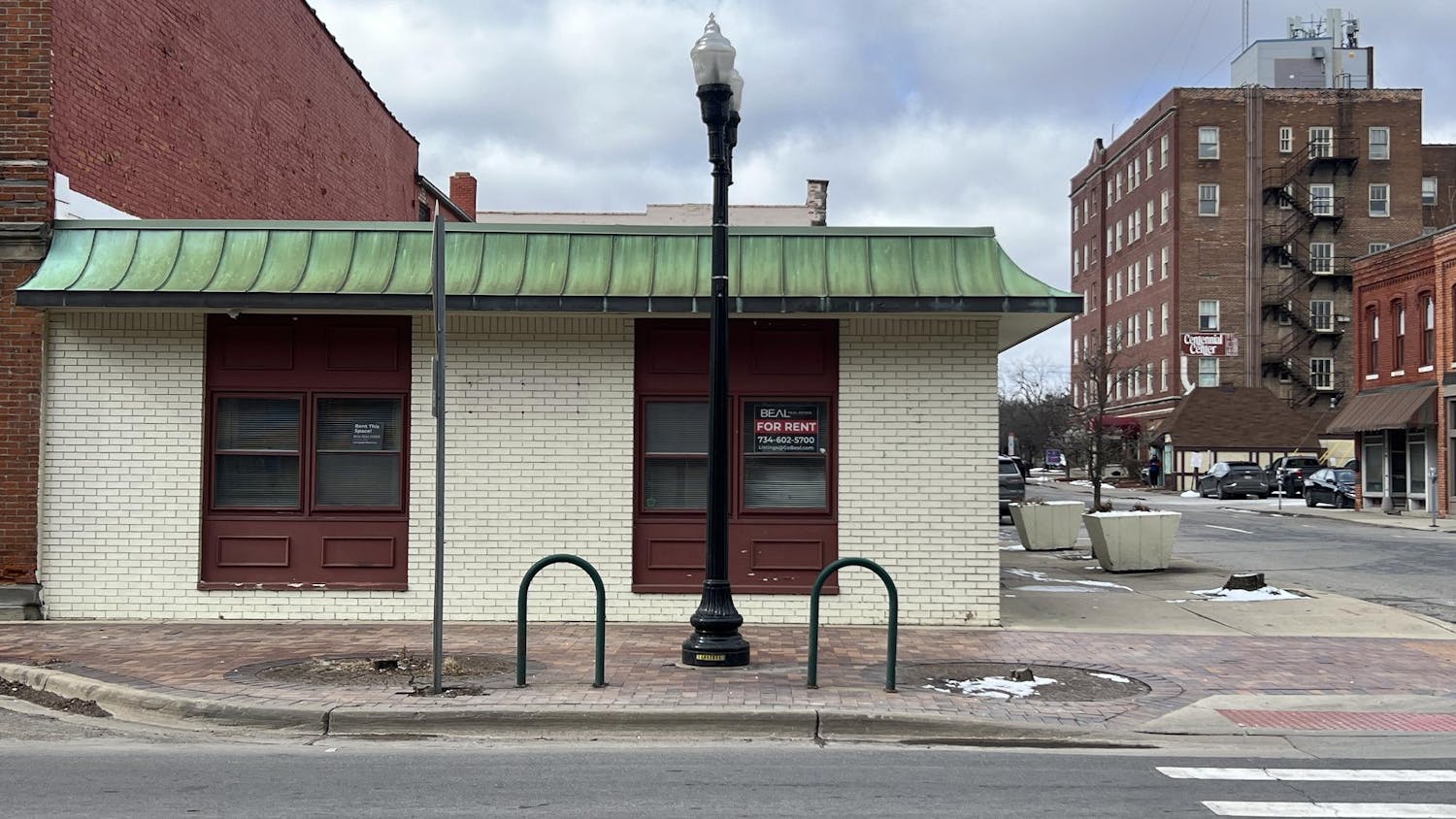 A white, brick, one-story building with red, wooden window frames. A sign in the window says that the building is available for rent.