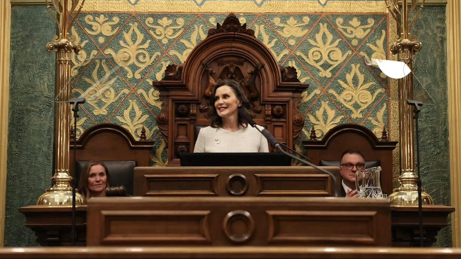 Gov. Whitmer stands at a wooden podium, backed by a green and gold wall. Sen. Winnie Brinks and Rep. Matt Hall sit behind her.