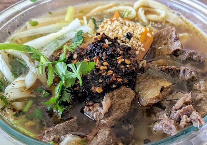 A ramen bowl that has beef chunks in the right bottom corner of the photo, and noodles in the top left side. The dish has chili paste and sliced spring onion as seasoning. 
