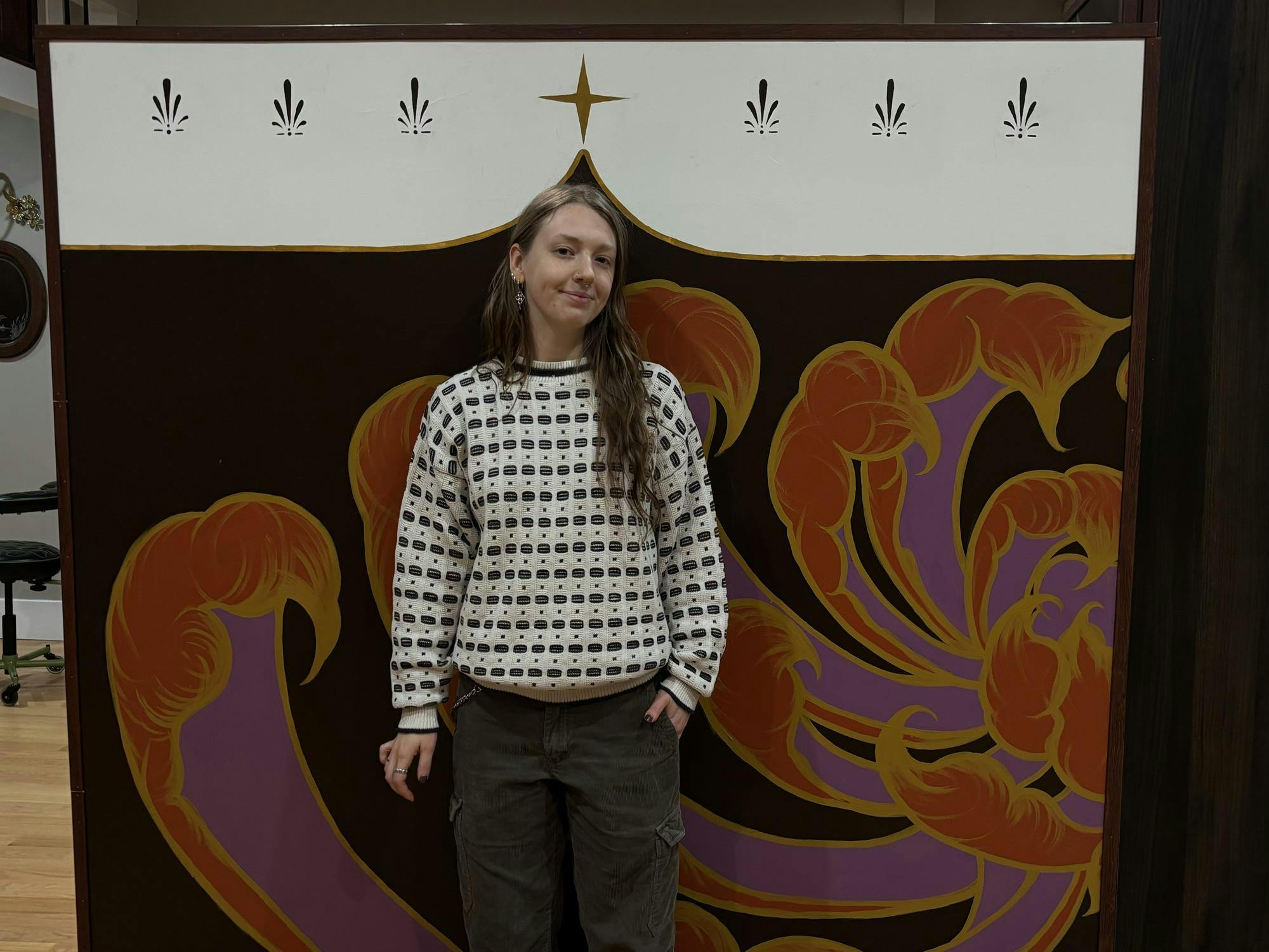 Rae Brooks-Pauli, wearing a white sweater and cargo pants, stands in front of a colorful backdrop and smiles softly.