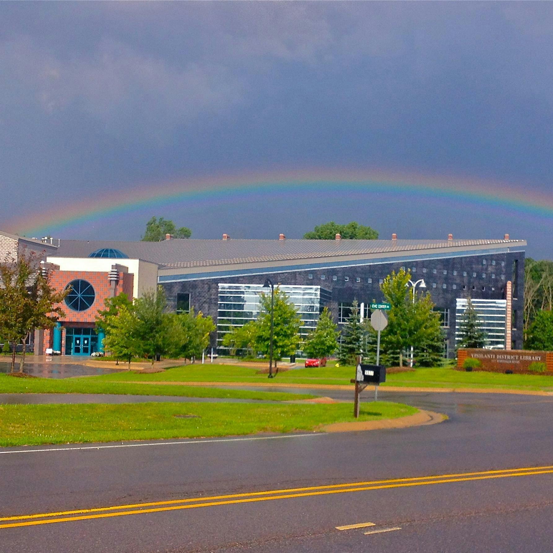 A building with a red-and-blue-brick entrance. Extending from the right side of the entrance is a long gray-colored section of the building. Several green trees frame the entrance to the building, and there is green grass in the foreground of the photo. Overhead, the sky is blue, and there is a rainbow visible over the building.