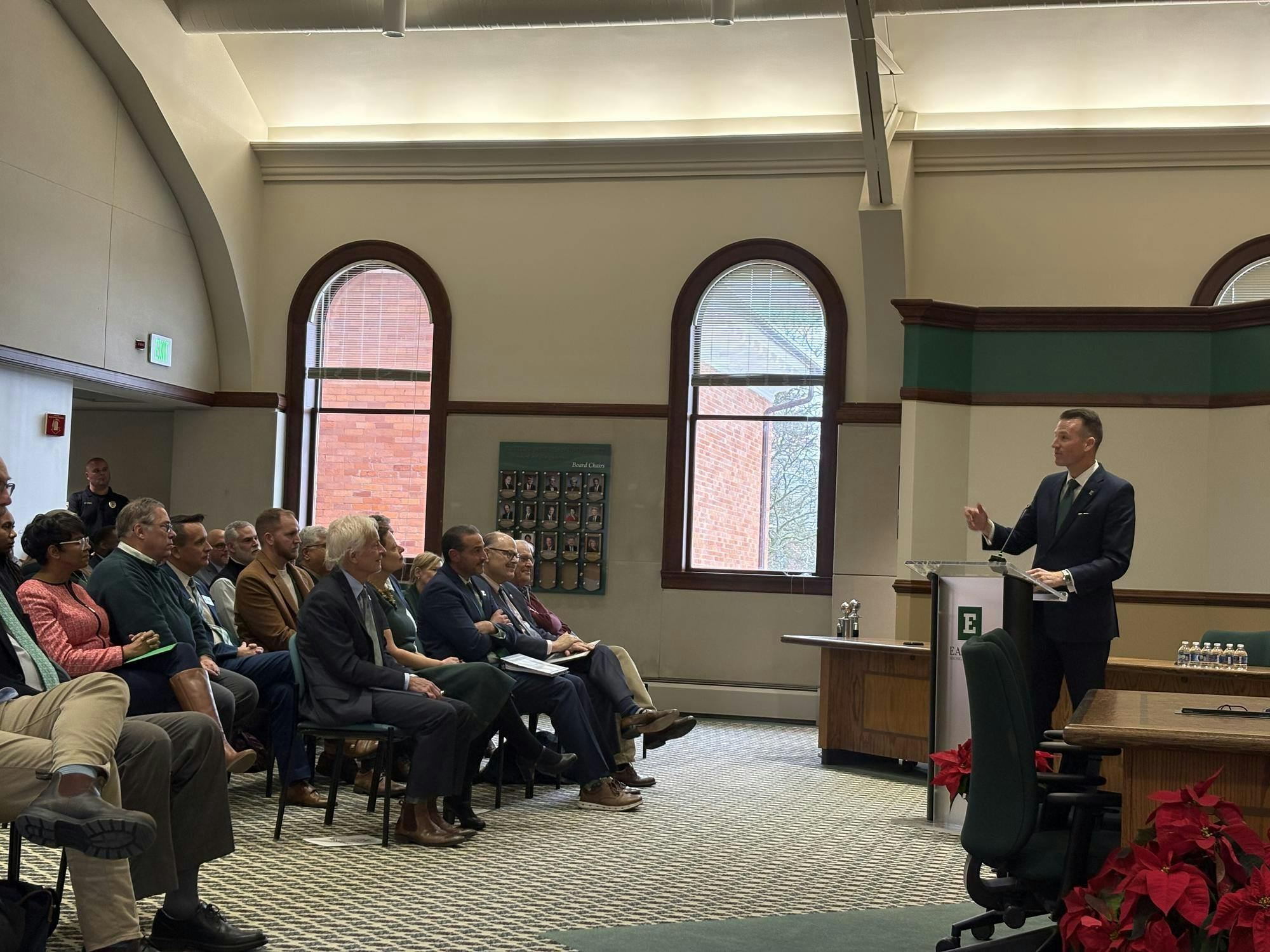 Brendan Kelly, dressed in a dark blue suit, speaks to a seated audience from behind an EMU-labeled podium.