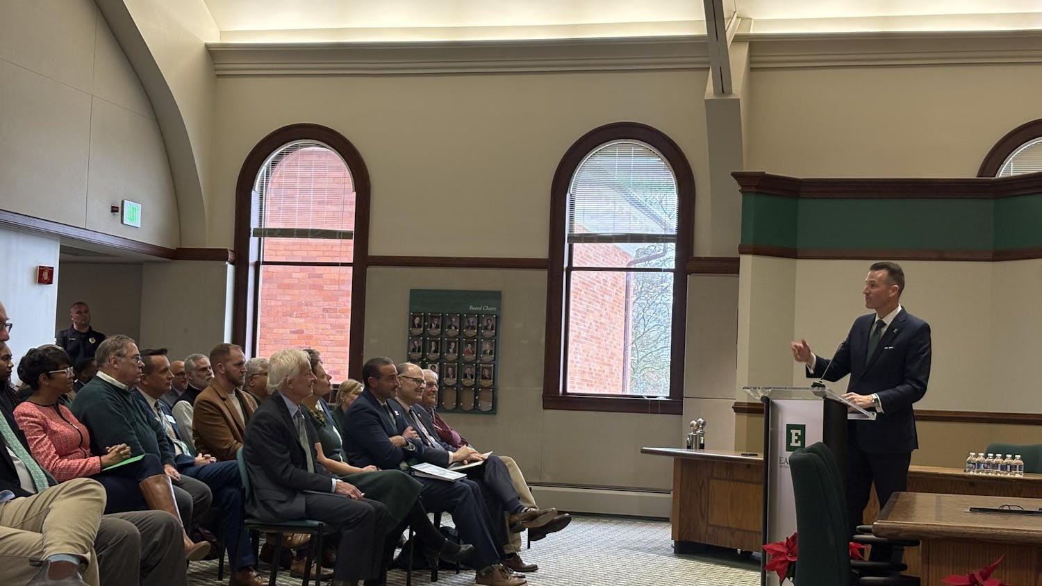 Brendan Kelly, dressed in a dark blue suit, speaks to a seated audience from behind an EMU-labeled podium.