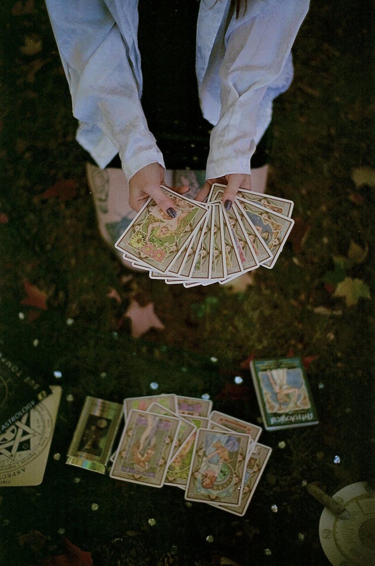 Close-up of a deck of tarot cards featuring soft, earth-tone pastels and feminine figures. The artist kneels on the ground outdoors in the background and a close-up of their hands holding the cards constitutes the foreground.