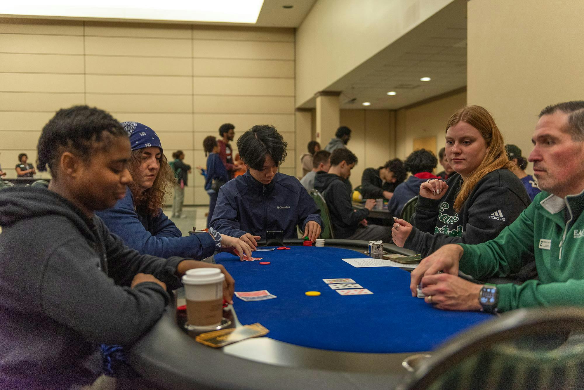 5 people sit around a table with a blue top, 3 of them are playing a card game while 2 of them are dealing the cards.
