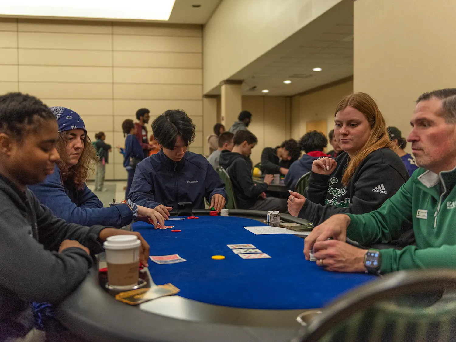 5 people sit around a table with a blue top, 3 of them are playing a card game while 2 of them are dealing the cards.