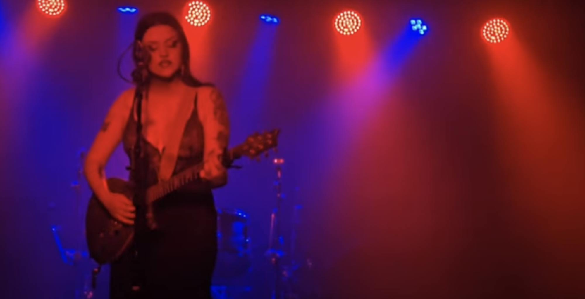 Dollie Rot plays on stage, holding an electric guitar and standing behind a microphone. Rot is surrounded by red and blue spotlights.