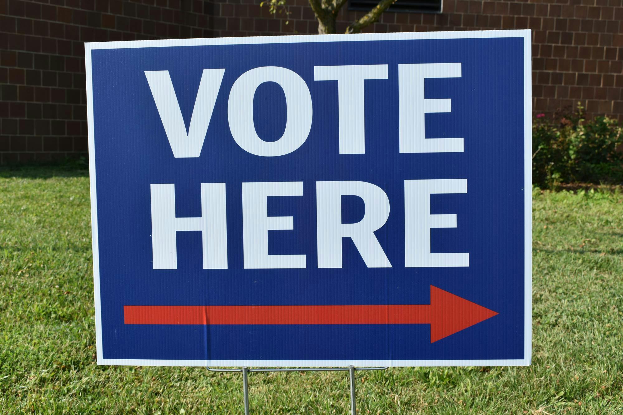 A blue and white sign that says "vote here" in white text written in all capital letters across two lines. On the third line underneath the text, there is a red arrow pointing to the right.

