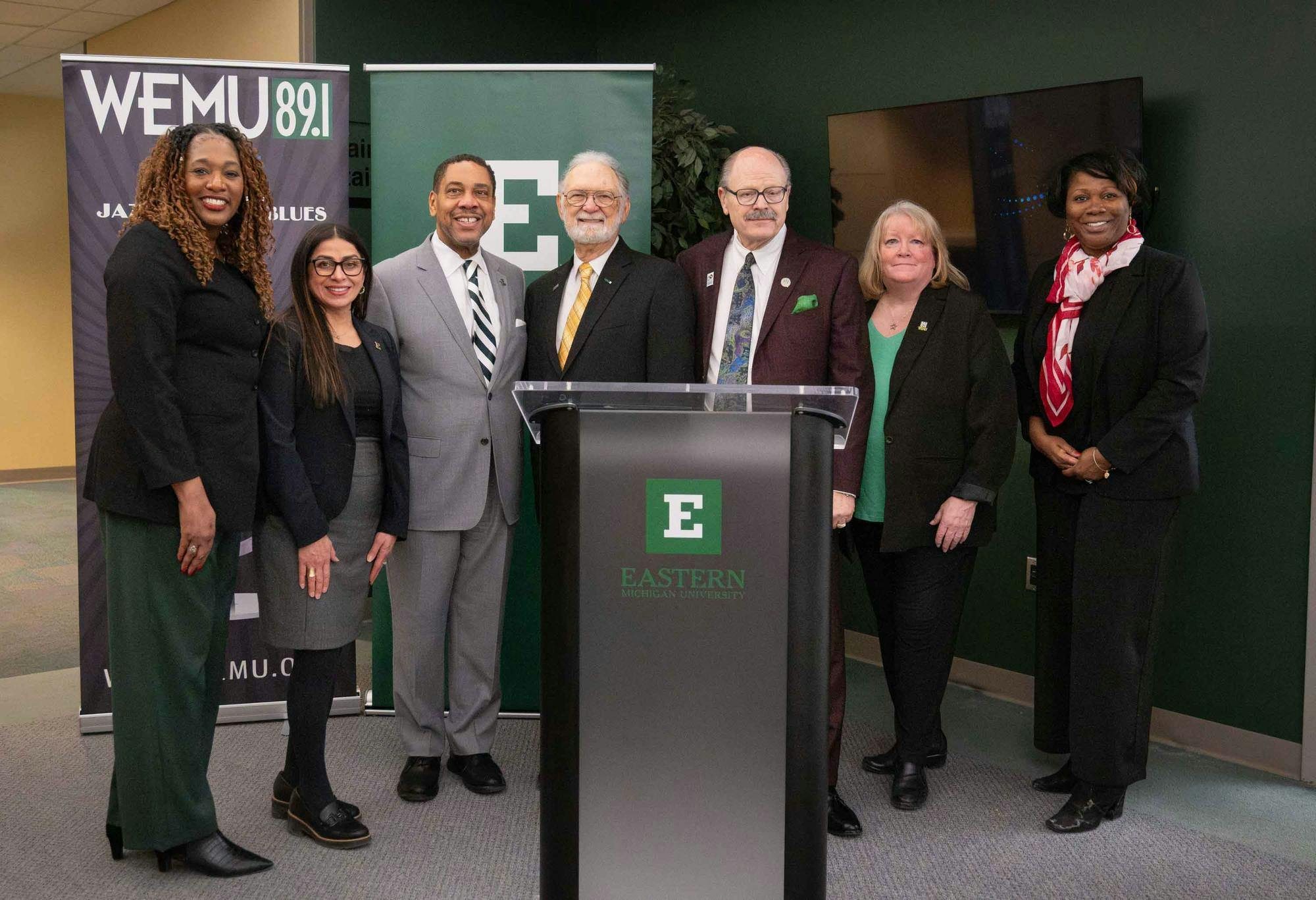 7 people stand near a podium and smile at the camera in front of WEMU and EMU branded banners.