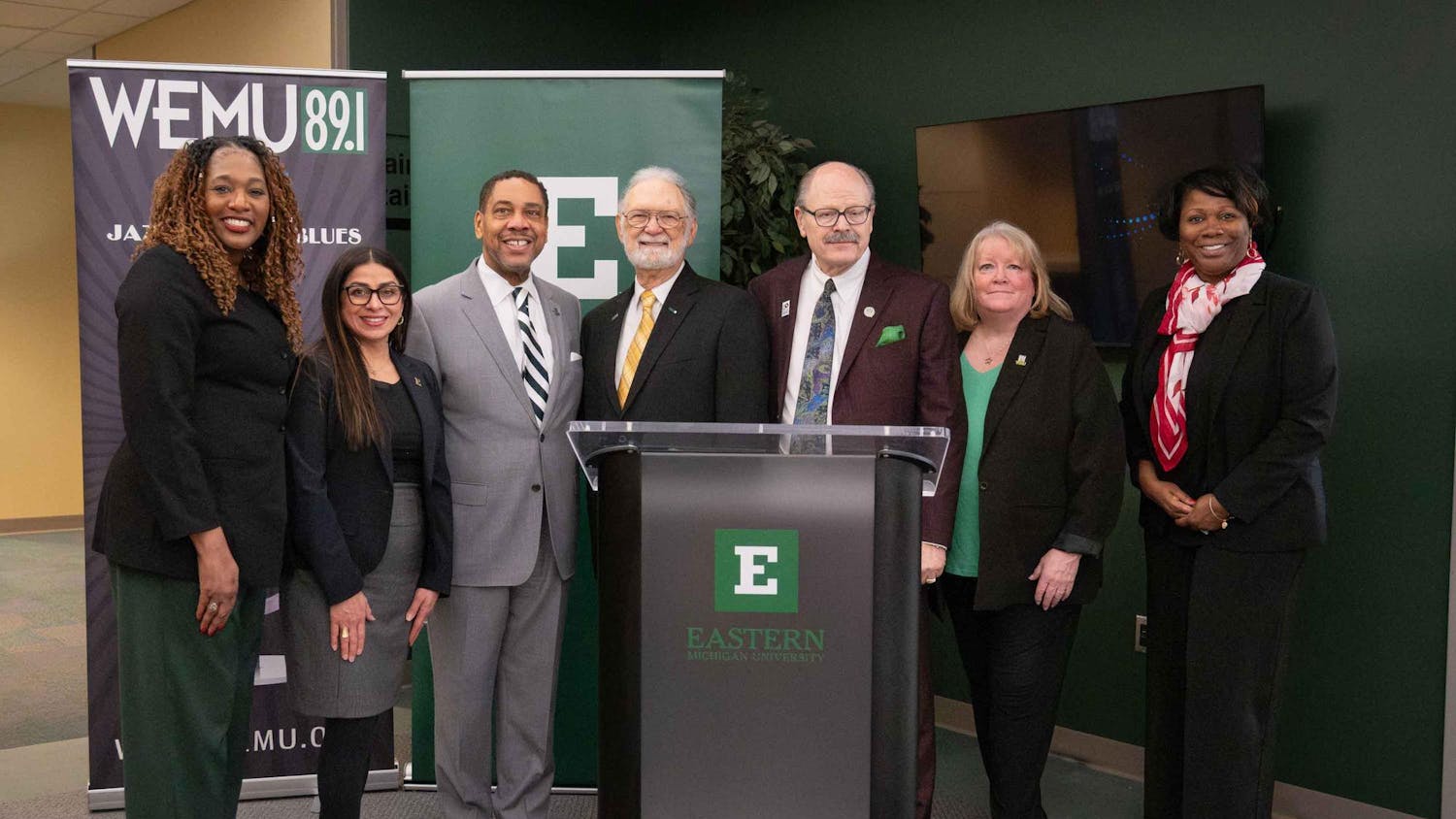 7 people stand near a podium and smile at the camera in front of WEMU and EMU branded banners.