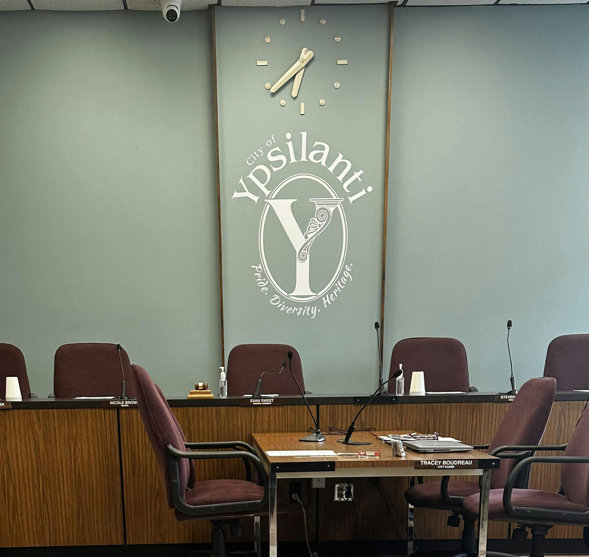Several chairs are sitting behind a brown desk with microphones on it. There is a teal blue wall with a clock. Underneath is white text that says "City of Ypsilanti", then a big Y in an oval, and under that is the text "Pride. Diversity. Heritage." 