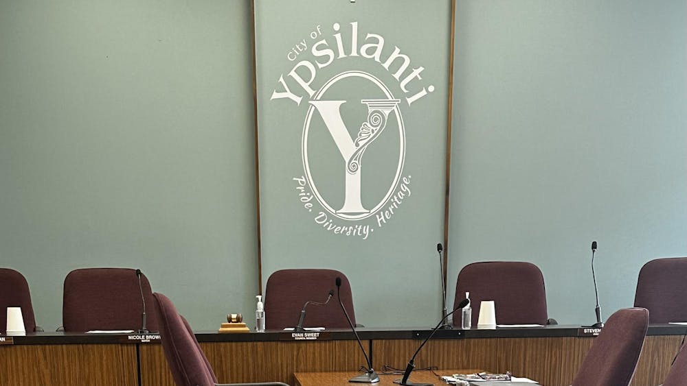 Several chairs are sitting behind a brown desk with microphones on it. There is a teal blue wall with a clock. Underneath is white text that says "City of Ypsilanti", then a big Y in an oval, and under that is the text "Pride. Diversity. Heritage."