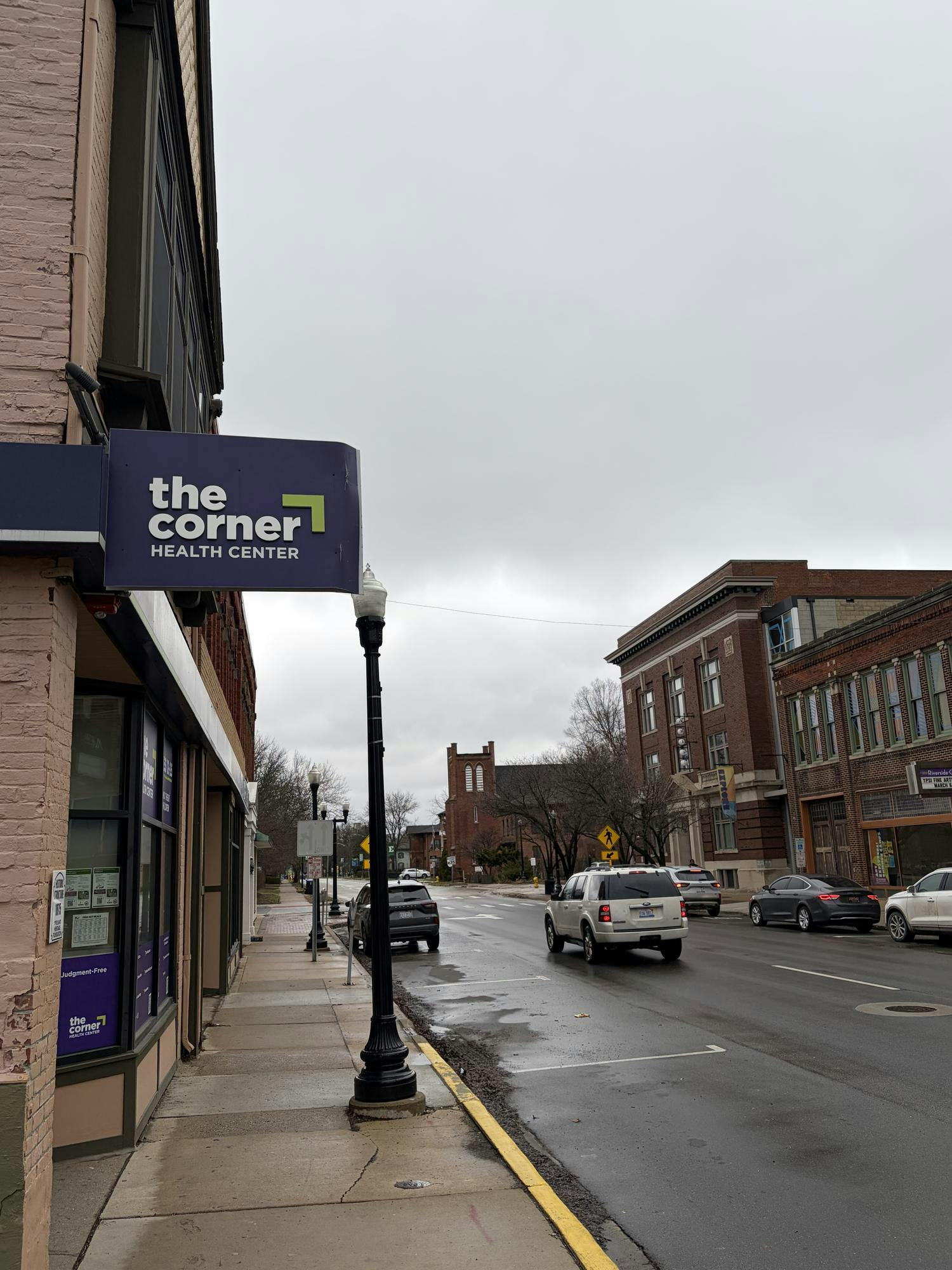 A sign for the Corner Health Center hangs from the side of a brick building along city street, with other brick buildings across the street and in the distance.