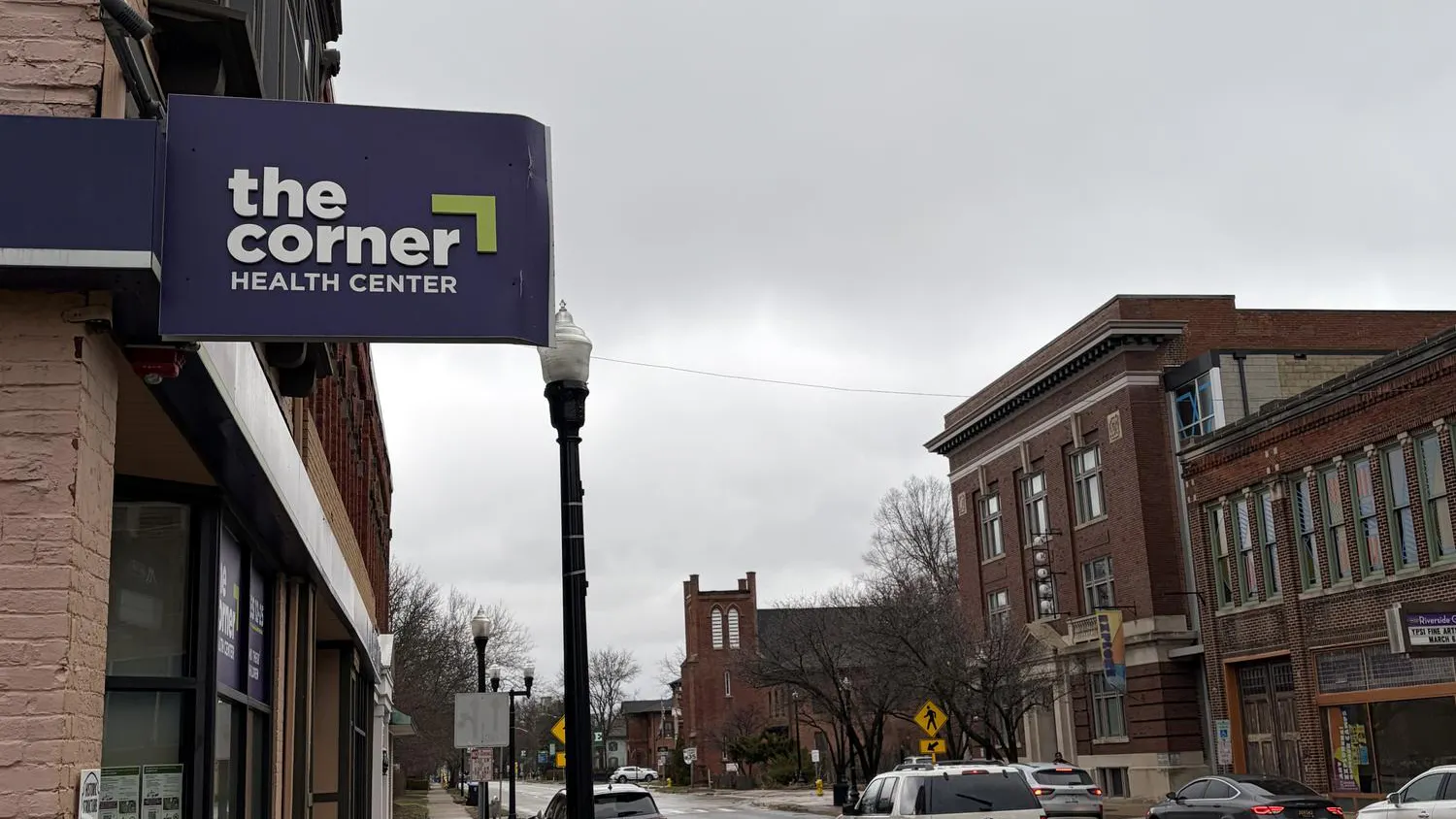 A sign for the Corner Health Center hangs from the side of a brick building along city street, with other brick buildings across the street and in the distance.