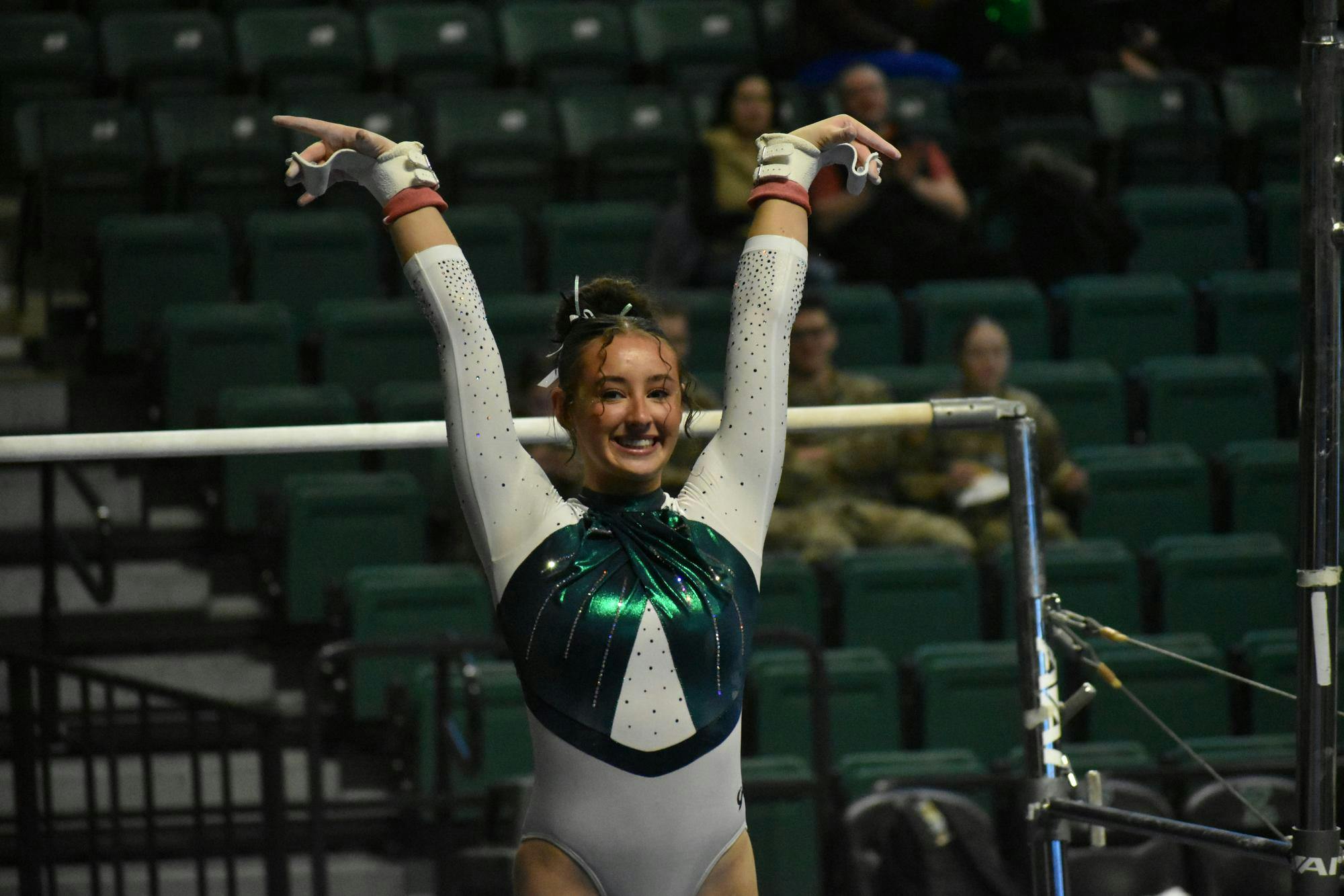 Gymnast Analiah Solorio salutes and smiles at the camera. The uneven bars she dismounted are in the background