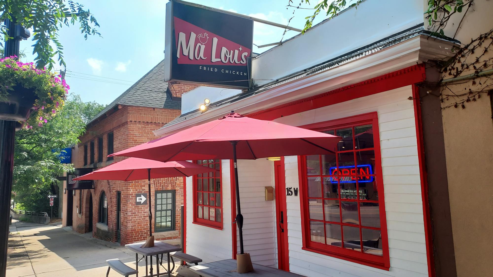 Ma Lou's entrance has two outdoor picnic tables shaded by big red umbrellas. The building's front has two glass windows with red trim with a door centered between them. In the rightmost window is an LED sign that reads "OPEN."