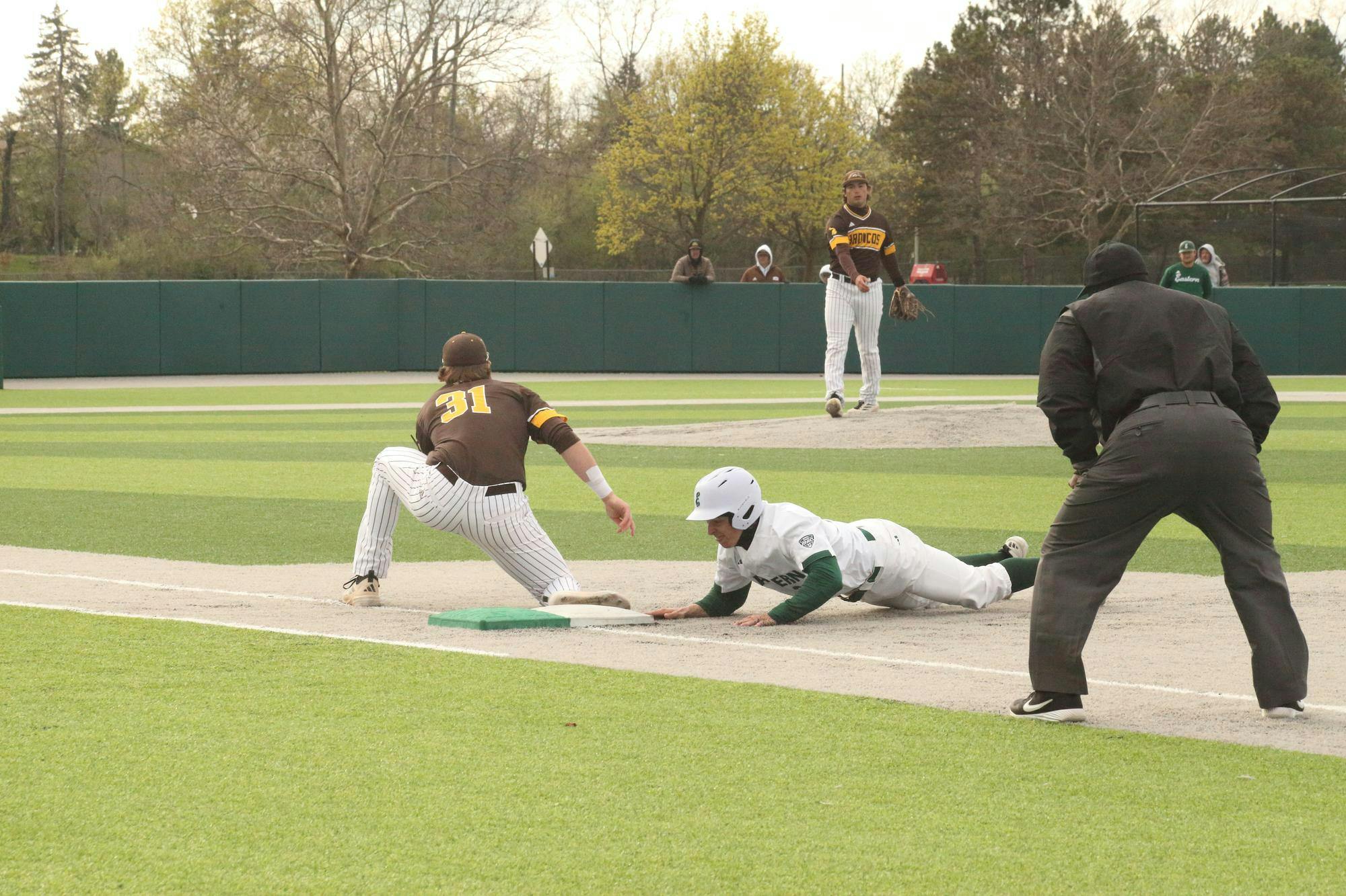 Baseball player slides face-first towards first base while the pitcher of the opposing team throws ball towards their teammate who is positioned near first base.