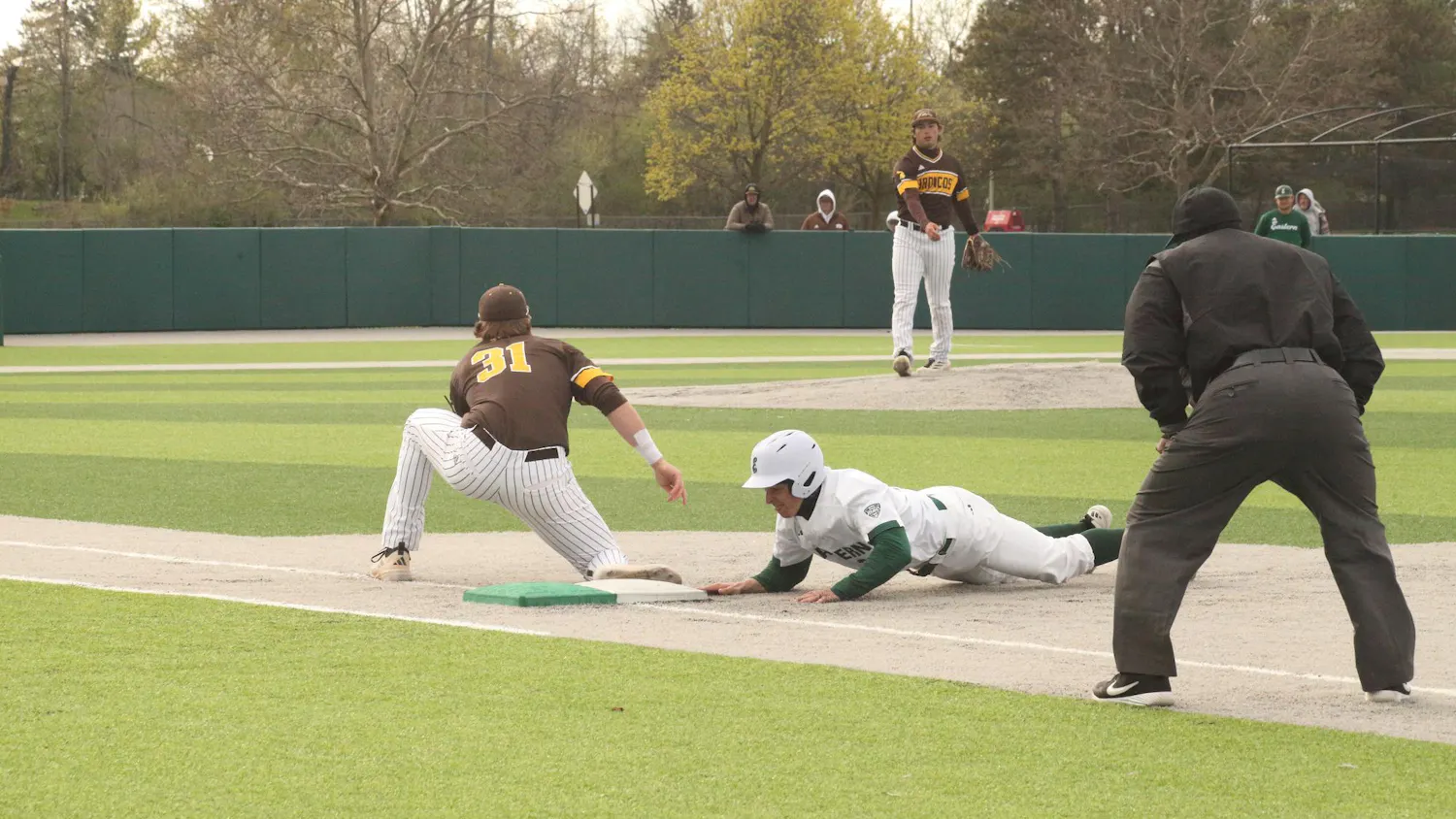 Baseball player slides face-first towards first base while the pitcher of the opposing team throws ball towards their teammate who is positioned near first base.