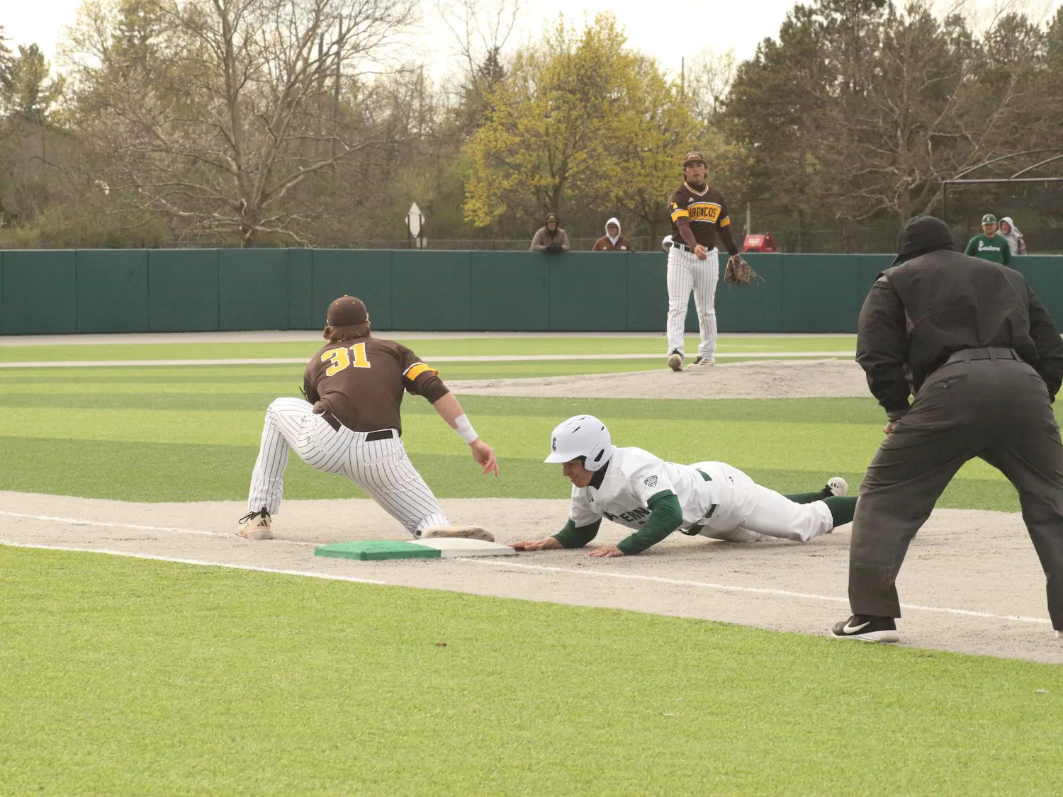 Baseball player slides face-first towards first base while the pitcher of the opposing team throws ball towards their teammate who is positioned near first base.