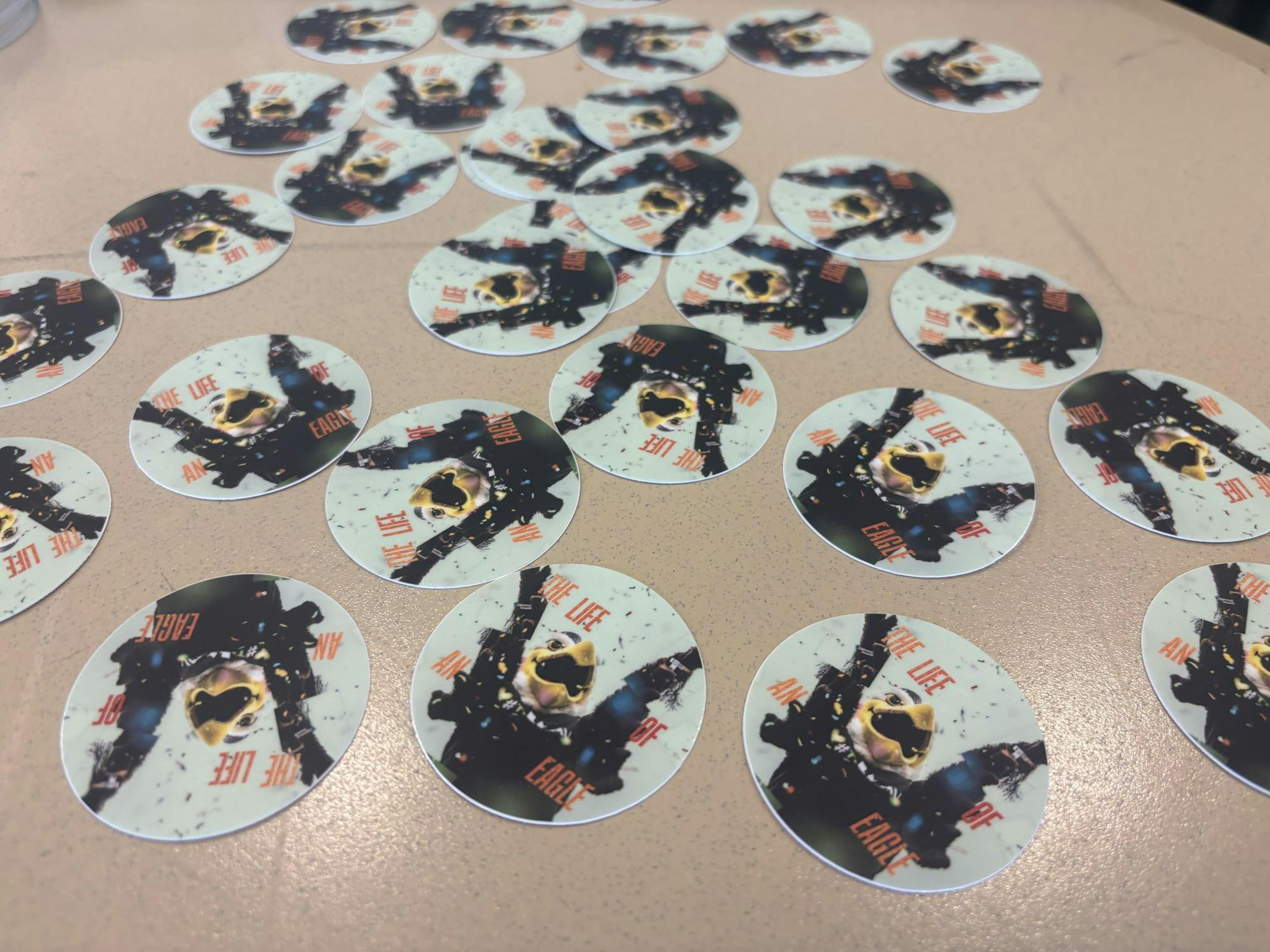 On a table, round stickers with a mint green background show an image of the EMU eagle mascot, Swoop. Stickers read "The Life of An Eagle" in glittery orange font.