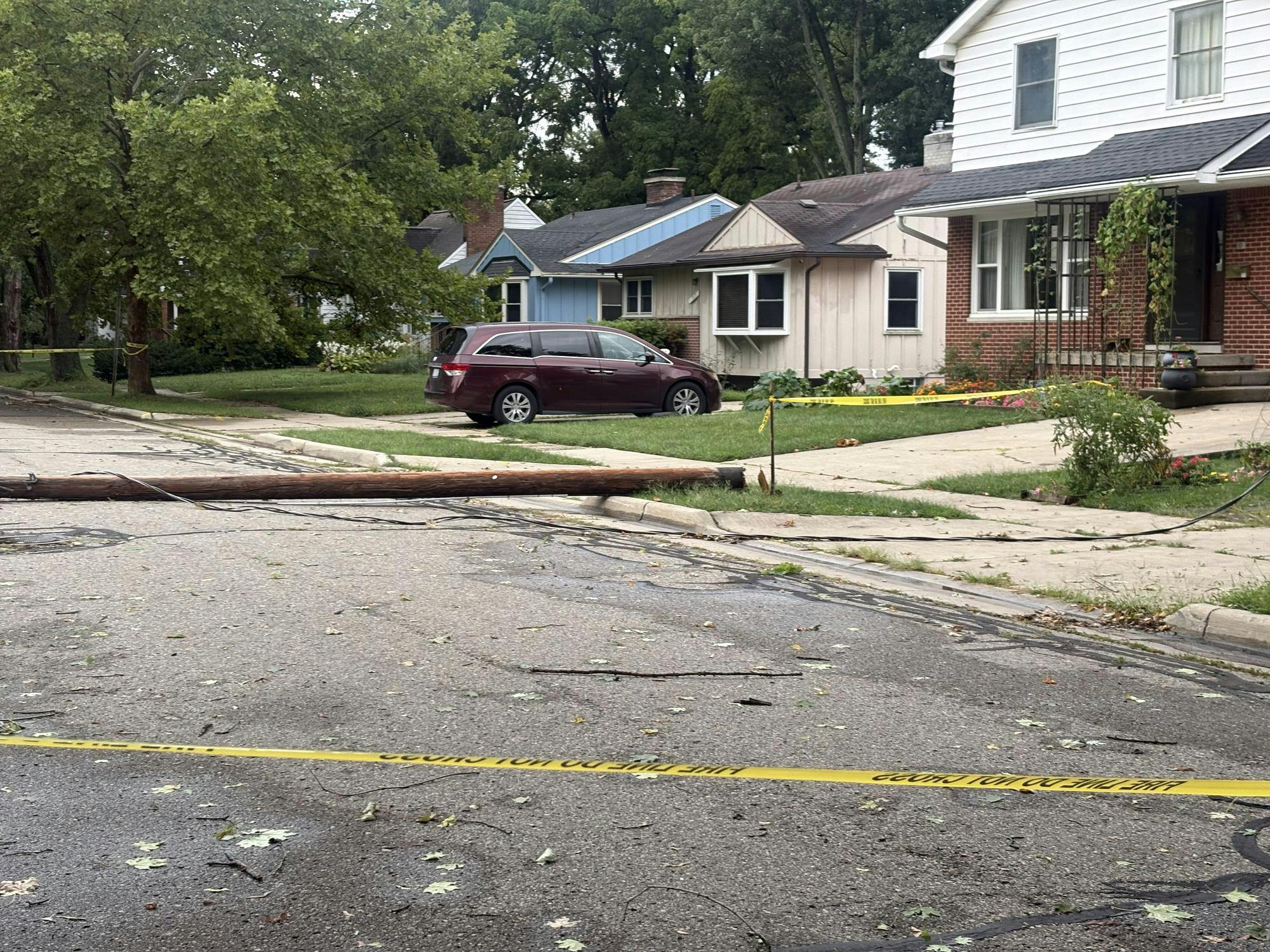 Yellow tape blocks off a road where a wooden electrical pole and sagging power lines lay across the middle of the street.