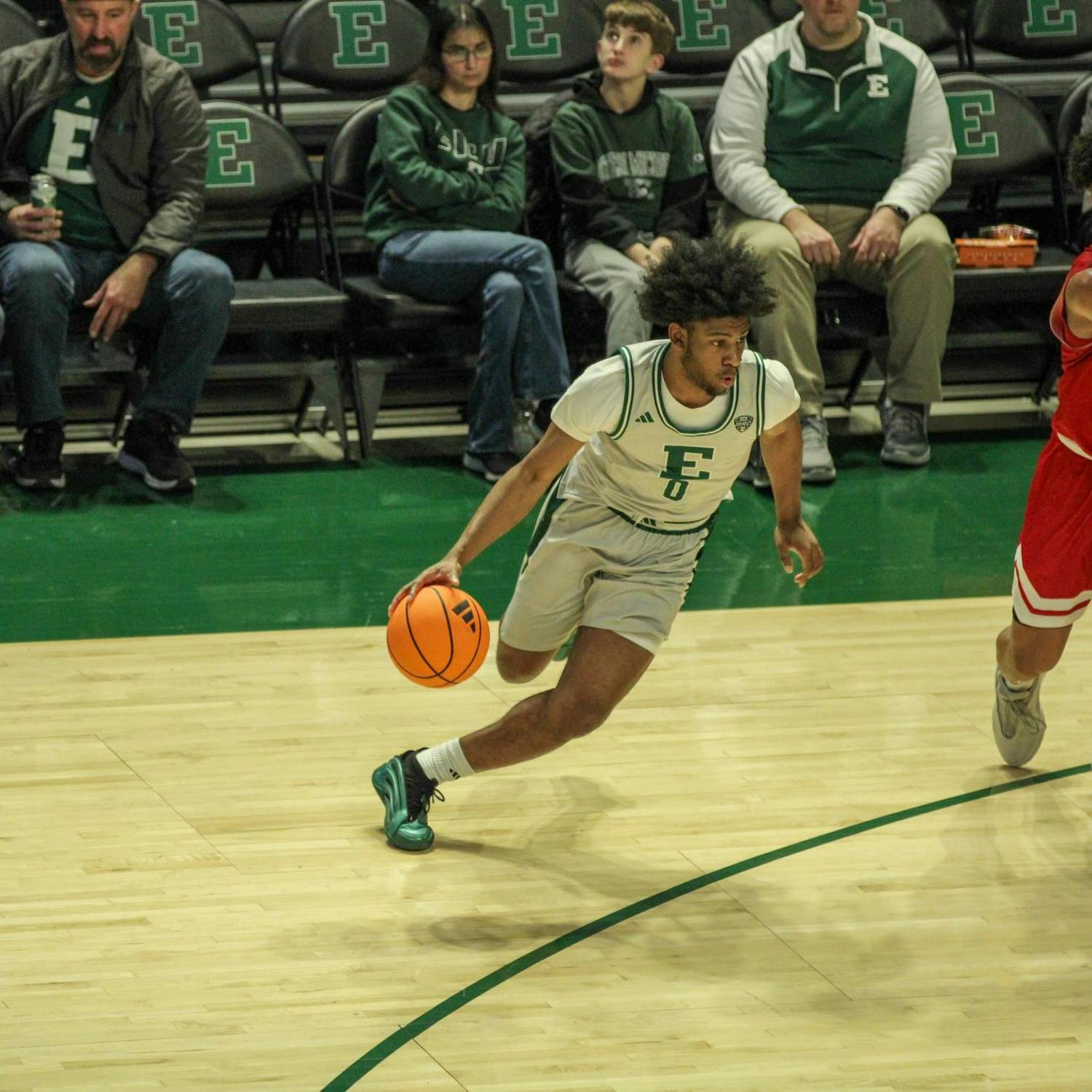 Image shows Eastern's No. 0 player Gregory Lawson II dribbling the ball as another player blocks the opponent. Fans watch from court side seats in the background.