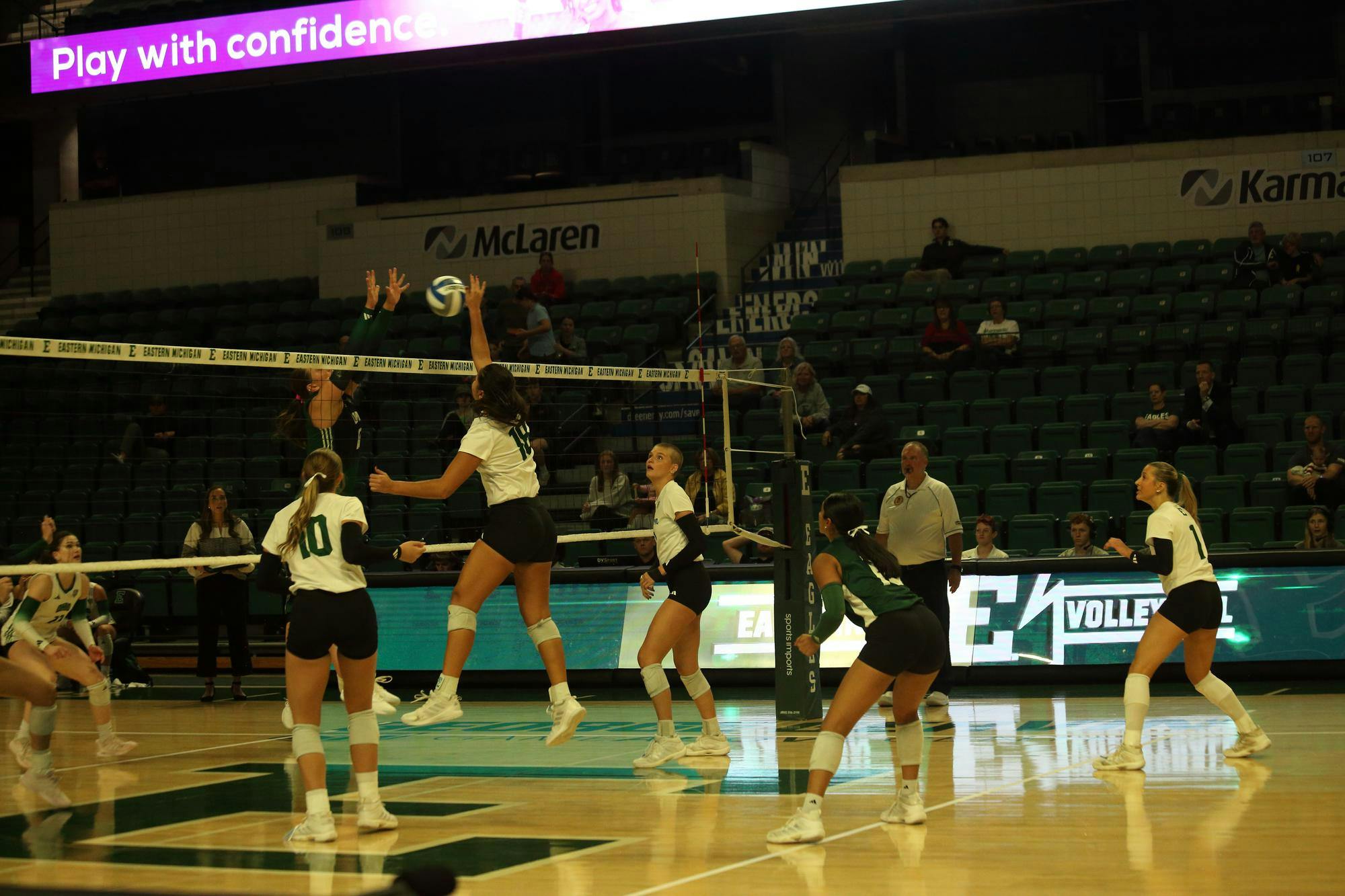 One player tips the ball over the net towards the other team while the other player leaps in the air; other players in uniform gather around on the court as the coach watches. A few spectators sit in the stands.